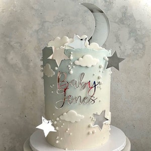 May include: A blue and white ombre cake decorated with white clouds, silver stars, and a silver crescent moon. The cake is topped with a silver mirror acrylic sign that says "Baby Jones".