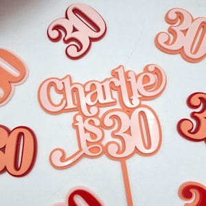 May include: A pink and red cake topper with the text "Charlie is 30" in a cursive font. The topper is made of acrylic and has a white stick for inserting into a cake.