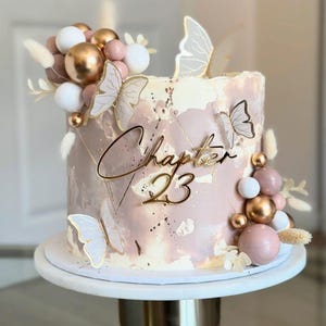 Op de afbeelding: Een versierde taart met roze en witte frosting, versierd met gouden en witte bollen, vlinders en de tekst "Chapter 23". De taart staat op een witte taartstandaard, perfect voor een verjaardag.