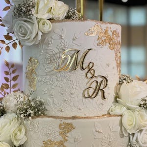 May include: A multi-tiered white wedding cake with detailed floral patterns and gold highlights. The cake is decorated with white roses and the gold letters "M&R". The cake has a textured surface and is decorated with gold leaf.