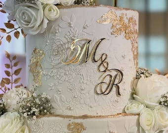 Puede incluir: Una tarta de boda blanca de varios niveles con intrincados diseños florales y detalles dorados. La tarta está adornada con rosas blancas y las letras doradas "M&R". La tarta tiene una superficie texturizada y está decorada con pan de oro.