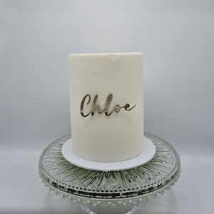 Pode incluir: Bolo branco com acabamento de espelho dourado e o nome "Chloe" escrito em letras douradas no topo. O bolo está em um suporte para bolo de vidro transparente.