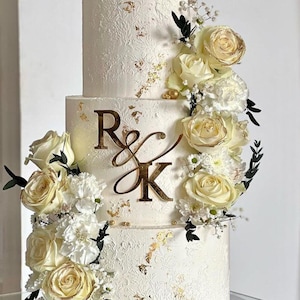 A three tier cream coloured cake decorated with white roses. On the front of the middle tier is a mirror gold acrylic monogram charm with the letters "R&K".