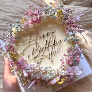 Può includere: Una torta quadrata color crema decorata con fiori colorati e le parole "Happy Birthday Afaf" in oro. La torta è adornata con fiori e nastri rosa, viola, gialli e blu.