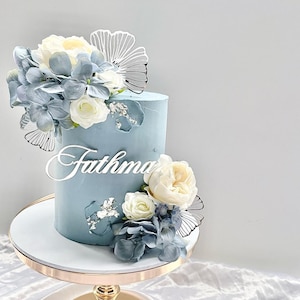 Peut inclure: Un gâteau bleu clair à deux étages décoré de fleurs blanches et bleues et d'un papillon argenté. Le gâteau est surmonté d'une pancarte en acrylique blanche qui dit "Fathma".