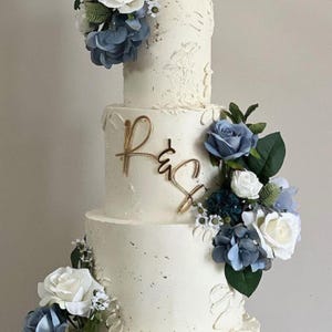 Op de afbeelding: Een witte bruidstaart met meerdere lagen, versierd met blauwe en witte bloemen en groene bladeren. De taart heeft goudkleurige initialen aan de zijkant. Een feesttaart voor een bruiloft of speciale gelegenheid.