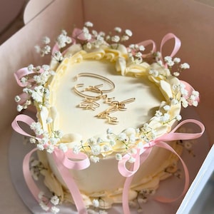 Può includere: Una torta rotonda, glassata bianca, decorata con fiori bianchi, nastri rosa e decorazioni dorate. La torta è in una scatola bianca, pronta per essere regalata o servita. La torta è un dolce per un'occasione speciale.