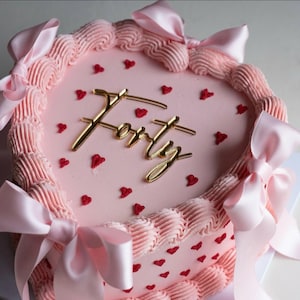 May include: A pink cake decorated with red heart sprinkles and a gold "Forty" topper. The cake features pink frosting swirls and is tied with pink satin bows, creating a celebratory design, perfect for a birthday.