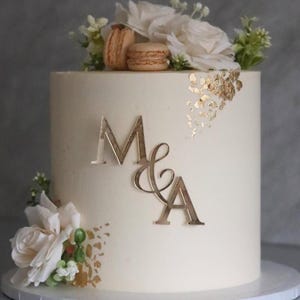 Può includere: Una torta rotonda color crema decorata con fiori bianchi, dettagli in foglia d'oro e due macarons. La torta è adornata con le iniziali dorate "M & A". Una torta per un matrimonio o un'occasione speciale.