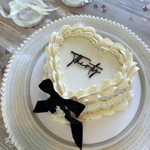 Pode incluir: Um bolo em forma de coração com cobertura branca e um laço preto. O bolo é decorado com cobertura creme nas bordas e a palavra "Thirty" em escrita preta. O bolo está sobre um prato branco e um carregador branco decorativo.