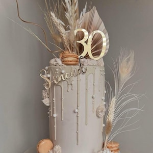 Peut inclure: Un gâteau à plusieurs étages avec un glaçage beige clair, orné de perles blanches et d'un cake topper doré "30". Le gâteau est décoré de fleurs séchées, de plumes et de macarons. Le nom "Sinead" est écrit en lettres dorées.