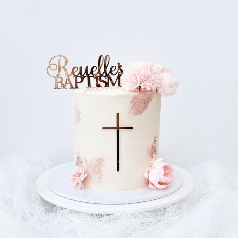 Mirror gold acrylic name topper with a spike stuck into the top of a cake. The topper has the name in a script font with "BAPTISM" written in a block font underneath. There is a mirror gold acrylic crucifix cake charm stuck on the front of the cake.