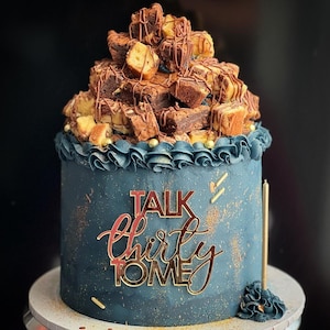Op de afbeelding: Een donkerblauwe cake met een gegolfde frostingrand, gegarneerd met een stapel brownies en stukjes cake overgoten met chocolade. De cake heeft gouden accenten en een gouden "Talk Thirty To Me" topper en een gouden kaars.