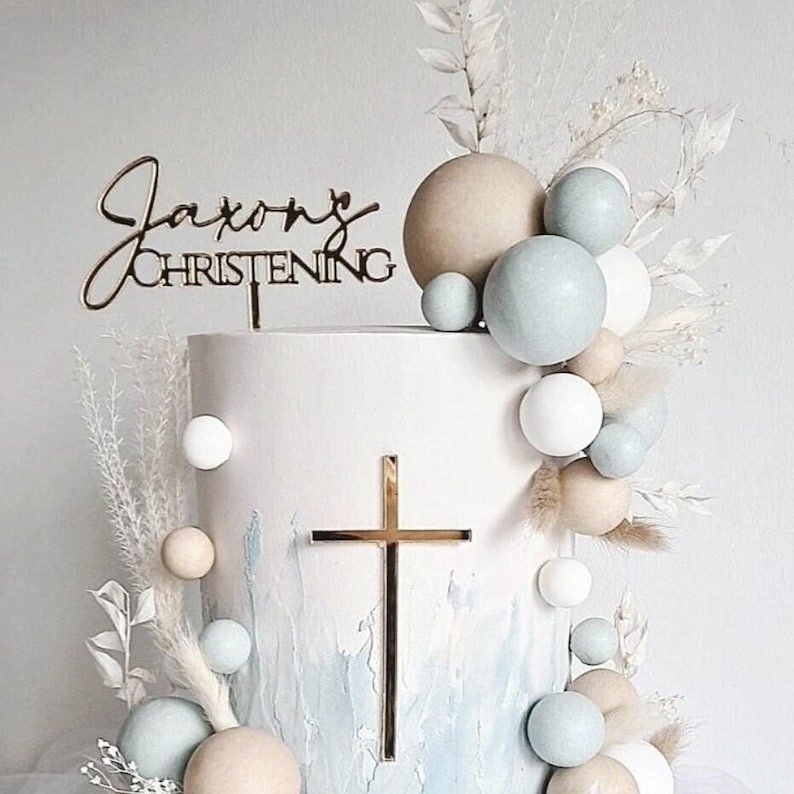 Mirror gold acrylic name topper with a spike stuck into the top of a cake. The topper has the name in a script font with "CHRISTENING" written in a block font underneath. There is a mirror gold acrylic crucifix cake charm stuck on the front of the cake.