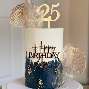 May include: A birthday cake with white and dark blue frosting, decorated with gold accents. The cake features a "25" topper and a "Happy Birthday Ayo" message. The cake is on a white surface.