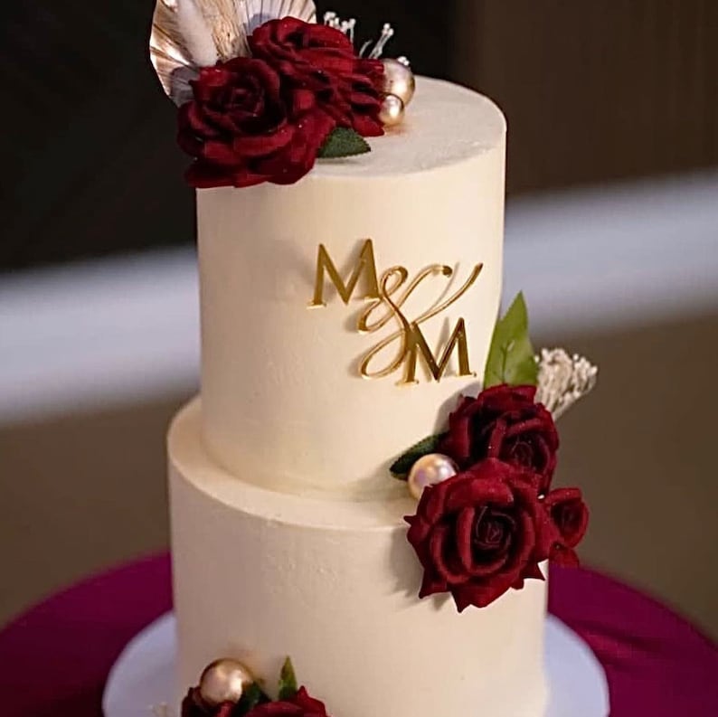 A two tier cream coloured cake decorated with red roses. On the front of the top tier is a mirror gold acrylic monogram charm with the letters "M&M".