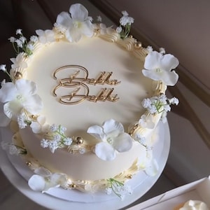 Peut inclure: Un gâteau rond de couleur crème décoré de fleurs blanches, de petites fleurs blanches et d'accents dorés. Le gâteau porte l'inscription dorée "Bella Dekki". Le gâteau est posé sur un présentoir à gâteau blanc.