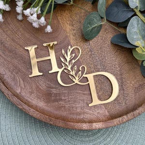 Op de afbeelding: Gouden acryl taarttopper voor een bruiloft met de letters "H & D" en een elegant bloemmotief. De topper staat op een houten dienblad met groen en witte bloemen op de achtergrond.