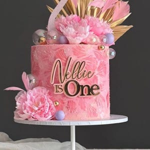 Puede incluir: Una tarta rosa glaseada con las palabras "Nellie is One" en letras doradas. La tarta está decorada con flores rosas, detalles dorados y esferas plateadas. La tarta está sobre un soporte blanco.
