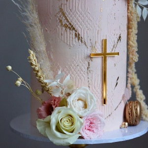 May include: A pink cake with gold accents and a gold cross on the side. The cake is decorated with white and pink flowers and wheat.