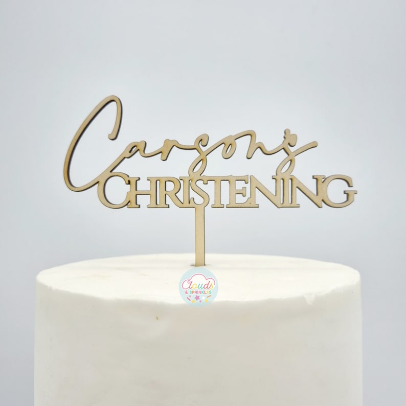 Wooden name topper with a spike stuck into the top of a cake. The topper has the name in a script font with "CHRISTENING" written in a block font underneath.