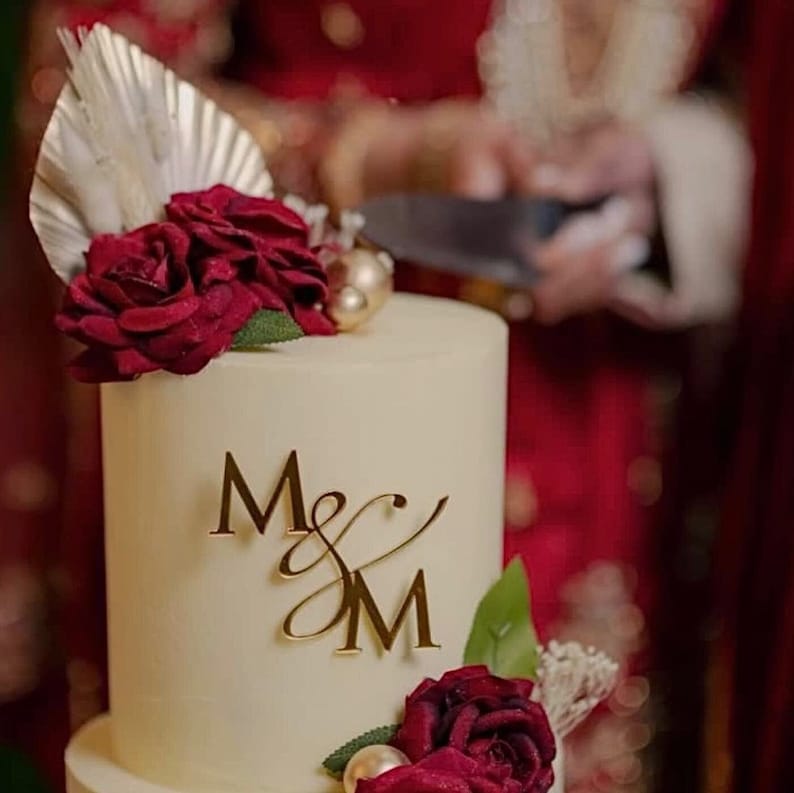 A two tier cream coloured cake decorated with red roses. On the front of the top tier is a mirror gold acrylic monogram charm with the letters "M&M".