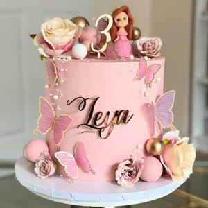 May include: A pink birthday cake decorated with pink and gold butterflies, roses, and a doll figurine. The cake has the name "Leya" in gold script and a gold number "3" on top. The cake sits on a white cake stand.
