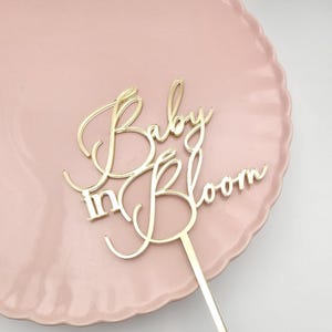 Może przedstawiać: Złoty topper na ciasto z napisem "Baby in Bloom" w ozdobnej czcionce. Topper umieszczony jest na różowym talerzu z falistym brzegiem. Elegancki design odpowiedni na baby shower lub uroczystość.