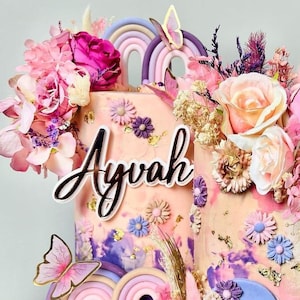 Puede incluir: Un pastel de dos pisos de color rosa decorado con flores, mariposas y un arcoíris. El pastel está cubierto con un letrero de acrílico blanco que dice "Ayvah".