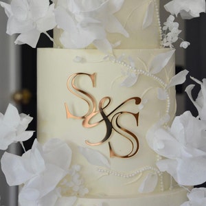Könnte beinhalten: Eine weiße Hochzeitstorte, verziert mit zarten Papierblumen und einem goldenen 'S & S' Monogramm.