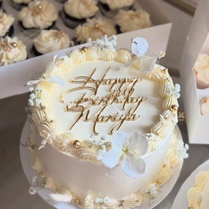 Puede incluir: Una tarta redonda de color crema con letras doradas que dicen "Happy Birthday Mariya". La tarta está decorada con glaseado blanco, chispas doradas y flores blancas. Cupcakes con glaseado blanco en el fondo.