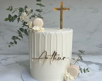 May include: White cake with white frosting and a gold cross topper. The cake is decorated with white chocolate drips and the name "Arthur" is written in gold script. The cake is surrounded by greenery.