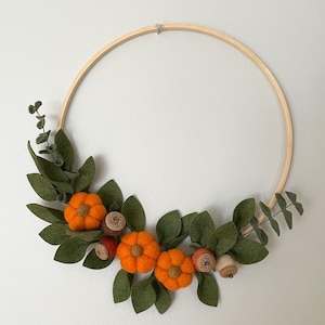Peut inclure: Une couronne sur le thème de l'automne avec un cerceau en bois orné de citrouilles en feutre, de feuilles vertes et de glands. Les citrouilles sont orange vif et les feuilles sont vert foncé. Les glands ont des chapeaux bruns.