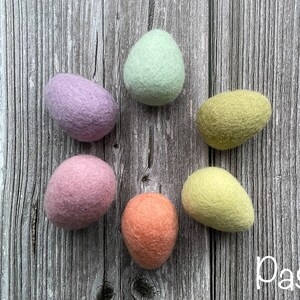 May include: Six pastel-colored felt eggs arranged in a circle on a wooden surface. The eggs are in shades of lavender, mint green, yellow-green, peach, and light pink. The word "Pastel" is written in white script in the lower right corner.