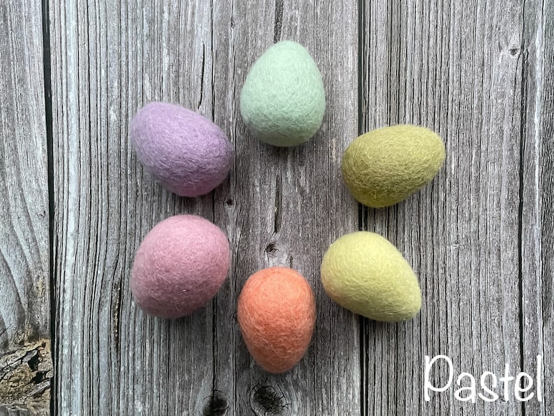 May include: Six pastel-colored felt eggs arranged in a circle on a wooden surface. The eggs are in shades of lavender, mint green, yellow-green, peach, and light pink. The word "Pastel" is written in white script in the lower right corner.
