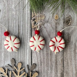 Puede incluir: Tres adornos navide&ntilde;os hechos a mano. Cada adorno es un c&iacute;rculo de fieltro blanco con rayas rojas, que se asemeja a un caramelo de menta. Tienen una cuenta roja y una cuerda para colgar. Los adornos se exhiben sobre una superficie de madera.