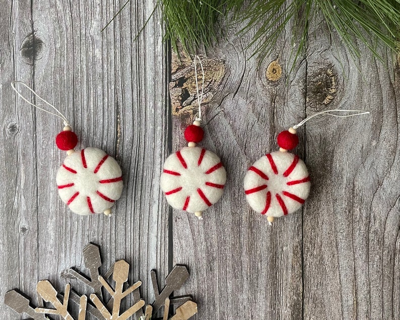 Puede incluir: Tres adornos navide&ntilde;os hechos a mano. Cada adorno es un c&iacute;rculo de fieltro blanco con rayas rojas, que se asemeja a un caramelo de menta. Tienen una cuenta roja y una cuerda para colgar. Los adornos se exhiben sobre una superficie de madera.