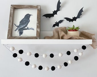 May include: A Halloween-themed shelf display featuring a framed watercolor painting of a crow, black bat cutouts, and a felt ball garland. The garland alternates black and white balls. A small succulent plant sits in a decorative pot.
