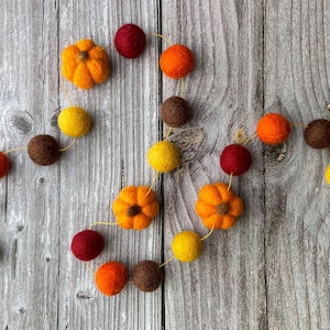 May include: A seasonal garland with felt pumpkins and balls in orange, yellow, brown, and red. The garland is strung on a thin gold thread, ideal for autumn decorations. It is displayed on a rustic wooden surface, perfect for fall decor.