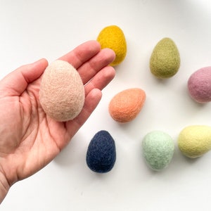 May include: A collection of handmade felt Easter eggs in various pastel colors, including pink, yellow, green, and blue. The eggs are arranged on a white surface, showcasing their soft texture and vibrant hues. These decorative eggs are perfect for Easter.