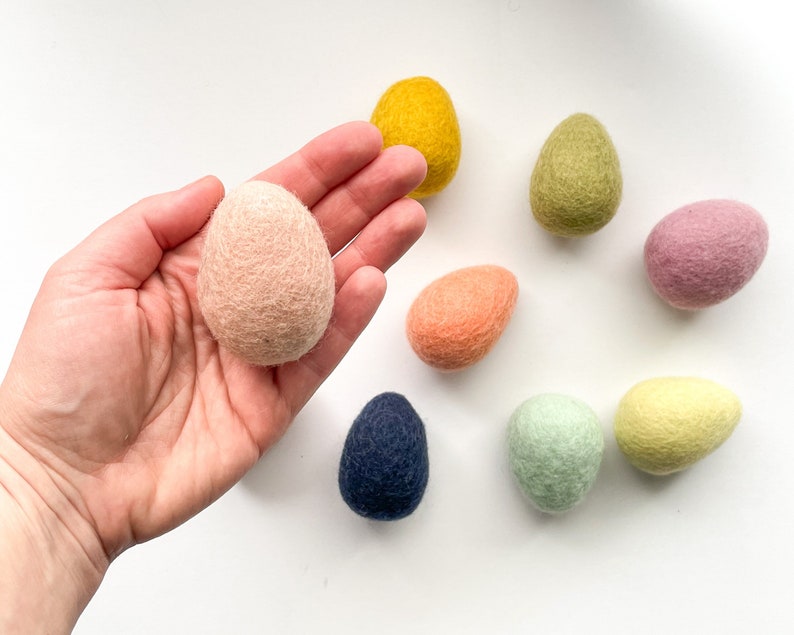 May include: A collection of handmade felt Easter eggs in various pastel colors, including pink, yellow, green, and blue. The eggs are arranged on a white surface, showcasing their soft texture and vibrant hues. These decorative eggs are perfect for Easter.
