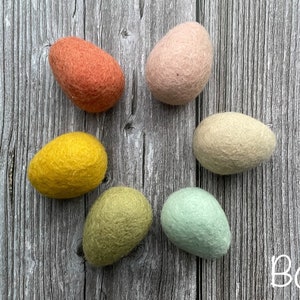 May include: Six felt eggs in shades of orange, pink, yellow, green, and cream are arranged on a wooden surface. The word "Boho" is written in white script in the lower right corner. These decorative eggs are perfect for Easter or spring decor.