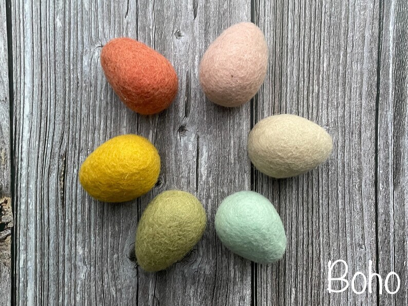 May include: Six felt eggs in shades of orange, pink, yellow, green, and cream are arranged on a wooden surface. The word "Boho" is written in white script in the lower right corner. These decorative eggs are perfect for Easter or spring decor.