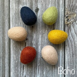 May include: Six felted egg-shaped ornaments in various colors: navy, light green, yellow, peach, rust, and light peach. The ornaments are arranged on a weathered wooden surface. The text "Boho 2" is in the bottom right corner.