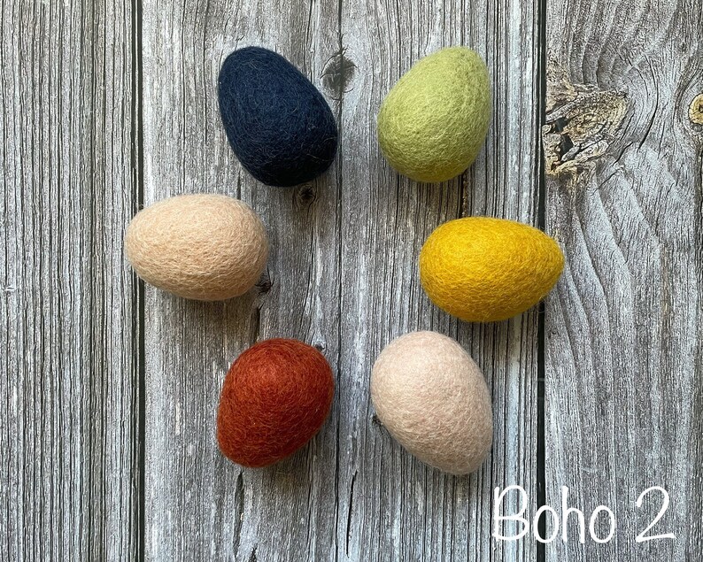 May include: Six felted egg-shaped ornaments in various colors: navy, light green, yellow, peach, rust, and light peach. The ornaments are arranged on a weathered wooden surface. The text "Boho 2" is in the bottom right corner.