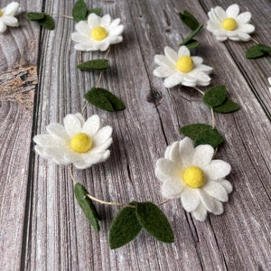 以下が含まれることがあります： 白いフェルトの花、黄色の中心、緑のフェルトの葉が付いたデイジーガーランド。 ガーランドは木製の表面に配置され、自然で気まぐれな美学を作り出しています。 春または夏の装飾に最適です。