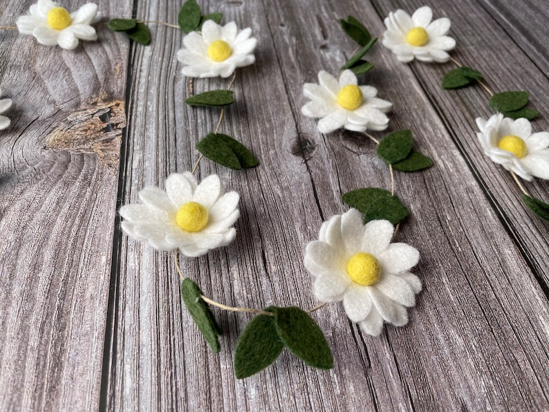 以下が含まれることがあります： 白いフェルトの花、黄色の中心、緑のフェルトの葉が付いたデイジーガーランド。 ガーランドは木製の表面に配置され、自然で気まぐれな美学を作り出しています。 春または夏の装飾に最適です。
