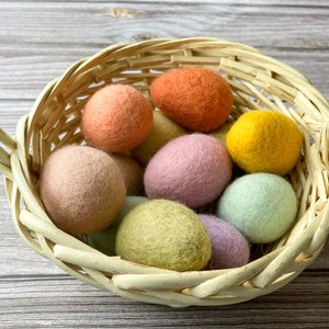 May include: A woven basket filled with colorful felt balls in shades of peach, orange, yellow, pink, green, and lavender. The basket has a handle and is made of light-colored woven material. The balls are round and have a soft, textured appearance.