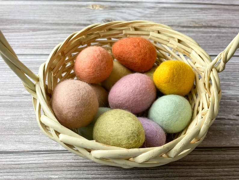 May include: A woven basket filled with colorful felt balls in shades of peach, orange, yellow, pink, green, and lavender. The basket has a handle and is made of light-colored woven material. The balls are round and have a soft, textured appearance.