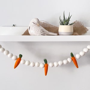 May include: A white shelf displays a ceramic bird, a small potted plant, and a burlap accent. A garland of white felt balls with orange carrot accents hangs below, creating a spring or Easter theme.